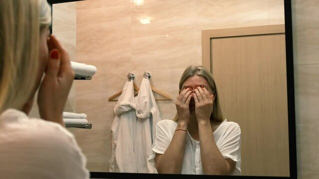Ageing. Sagging skin. A young woman does facelift exercises by looking in the bathroom mirror. Facebuilding. Modern care, relaxation of facial muscles. Facelift, plastic surgery