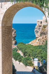 Beautiful view of Polignano a Mare with white stone houses, cliff, beach, blue sea and bathers surrounded by Mediterranean nature, vertical