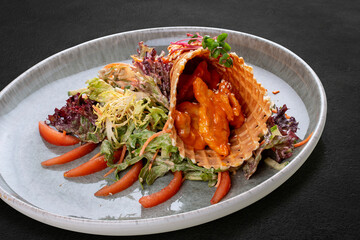 Salad with sweet and sour chicken in a waffle cone on a bed of lettuce leaves. On a dark background, isolated.