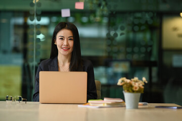 Confident female entrepreneur sitting in modern office with laptop computer and smiling at camera