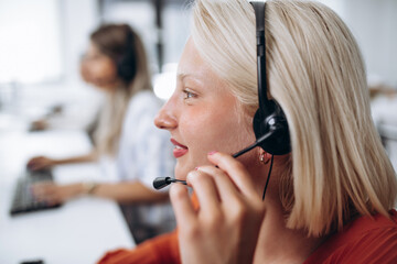 Beautiful smiling blond female call-center agent with headset working on support in the office.