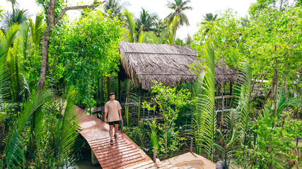 young man walking down pier near mekong delta river in Ben Tre Vietnam with straw roof hut in background surrounded by green jungle - obrazy, fototapety, plakaty