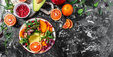 Healthy vegetarian buddha bowl salad with avocado, persimmon, blood orange, nuts, spinach, arugula and pomegranate on a dark background, Long banner format, top view