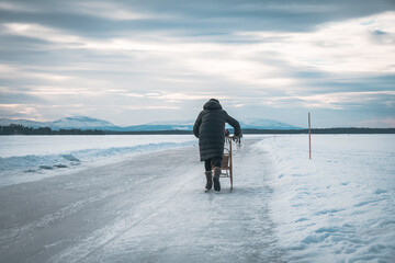 Elderly woman on ice skating track called "medvinden" on lake storsjön near Östersund (Sweden Lapland) riding sledge towards mountain view (Oviksfjällen).