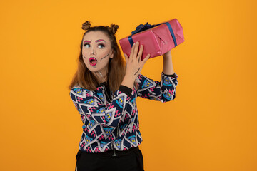 Portrait of young girl wearing creative makeup and shaking her gift box on orange background