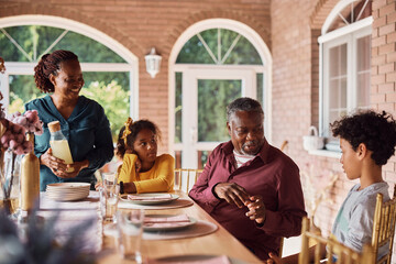 Happy African American kids and their grandparents talk at dining table on patio.