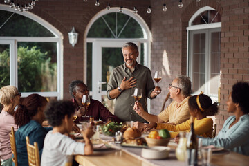 Happy mature man talks while toasting to his multiracial family during lunch at dining table.
