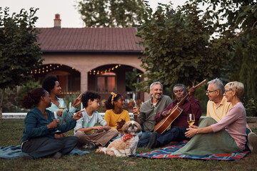 Senior black man plays acoustic guitar while relaxing with his multiracial family in backyard.