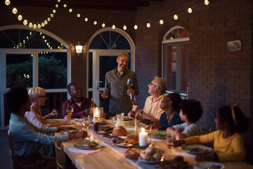 Happy man holding a toast while having dinner with his multiracial family on patio.