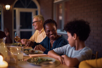 Happy black senior woman talks to her grandson during family dinner on patio.