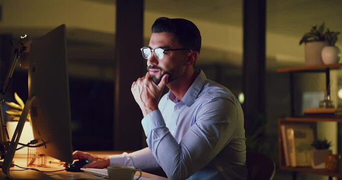 Young business man looking thoughtful while reading an email on a desktop computer and working late at night. One serious corporate male designer doing overtime to meet a deadline in a dark office