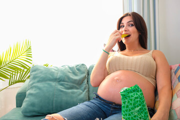 Pregnant young woman eating chips on the sofa at home. Food and cravings during pregnancy.