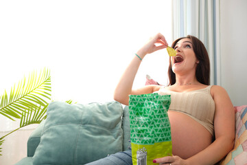 Pregnant young woman eating chips on the sofa at home. Food and cravings during pregnancy.
