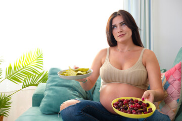 Pregnant young woman eating on the sofa at home. She is holding two plates in her hands and is...