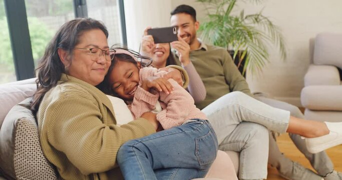 Adorable little girl playing with grandmother, laughing in home living room. Multi generation family enjoying quality time together. Smiling child being tickled while parents use a phone for photos
