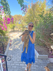 A young girl in a blue dress and sunglasses is holding a beautiful gray tabby cat in nature, Cyprus. A disgruntled cat tolerates human hands. The girl and the cat. Friends, animals and people