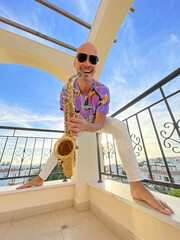 A young male saxophonist in white pants and a purple T-shirt stands on the balcony of a hotel in Cyprus and holds a saxophone musical instrument in his hands and smiles. Cheerful saxophonist