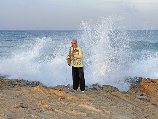 A young stylish bald male saxophonist stands against the backdrop of the sea and stormy high waves and plays the golden saxophone in Cyprus. Romantic Musician saxophonist