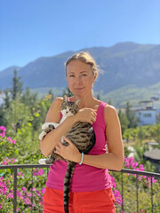 Young girl in summer holds in her arms, gently hugs young beautiful and fluffy gray-white cat. Cat in arms of girl, against background of beautiful pink flower, blue sky, mountains. Love animals