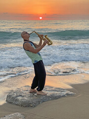 Young male saxophonist stands against background of sea and sunset, holds saxophone in his hands. Beautiful sunset on sea, sky. Musician saxophonist plays against background sea, sunset, silhouette