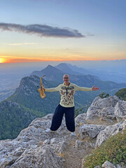 Young stylish bald male saxophonist stands on top of mountain against the backdrop of the sunset and the sun and plays the golden saxophone in Cyprus. Romantic Musician saxophonist. Above clouds