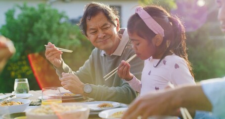 Proud grandfather watching his granddaughter eat while having a family lunch in a backyard. Young girl enjoying Asian food while learning to use traditional chopsticks with loving grandpa outdoors