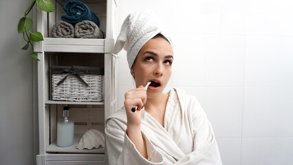 Cute young woman brushing her teeth in the bathroom, wearing a bathrobe and with her hair wrapped...