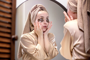 A well-groomed woman at home in a bathrobe and towel applies patches under the eyes from swelling...