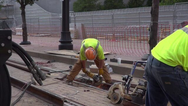 Slow motion video of a construction worker grinding the new steel railroad tracks in a city. Sparks fly in slow motion as another worker is in the foreground.