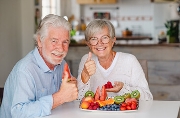 Happy beautiful retired senior couple having break with fresh seasonal fruit at home, thumb up looking at camera, healthy eating concept