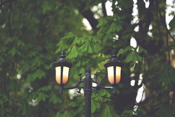 Old cast iron lamp post with two warm lanterns on trees background in evening. Concept of Nostalgia...
