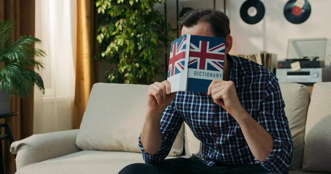 A man tries to memorize new English words. A bearded man sits on the couch and hits his head with a dictionary, hoping that he will finally remember the definition.