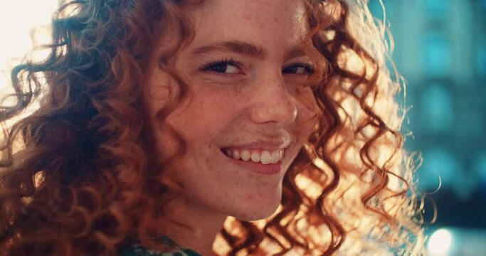 Close up face young sunshine young woman with red hair look at camera smile stand in the city streets. Beautiful town at sunlight. Feel happy. Slow motion