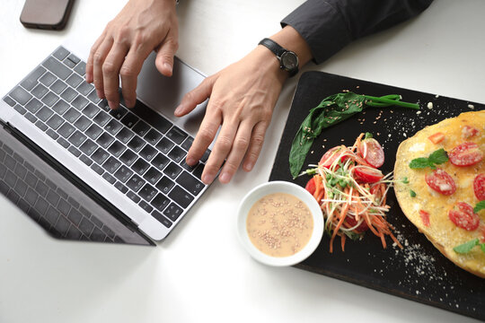 Top view of a wooden board with lunch in front of woman's hands, printing on the laptop. Lunchtime.