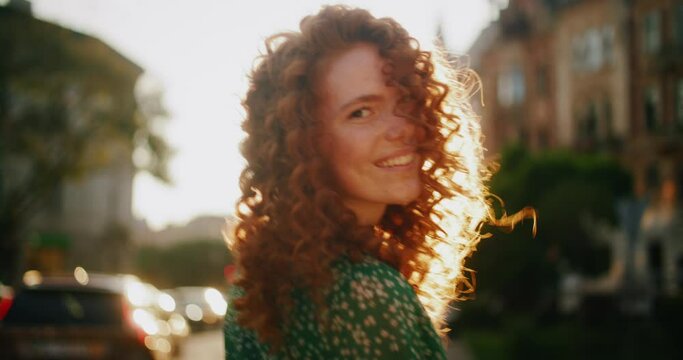 Happy young sunshine woman with red hair turning look at camera smile walking in the city streets. Beautiful town at sunlight. Portrait. Slow motion