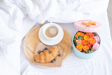 Obraz na płótnie Canvas Cup of coffee,croissant with fresh blueberries on wooden tray.Romantic breakfast in bed with gift box of orange roses,macarons.Birthday,Valentine's day morning. Honeymoon concept.Delivery service