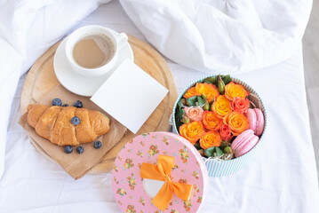 Cup of coffee,croissant,blueberries,empty note on wooden tray.Romantic breakfast in bed with gift box of orange roses,macaroons.Birthday,Valentine's day morning. Honeymoon concept.Delivery service