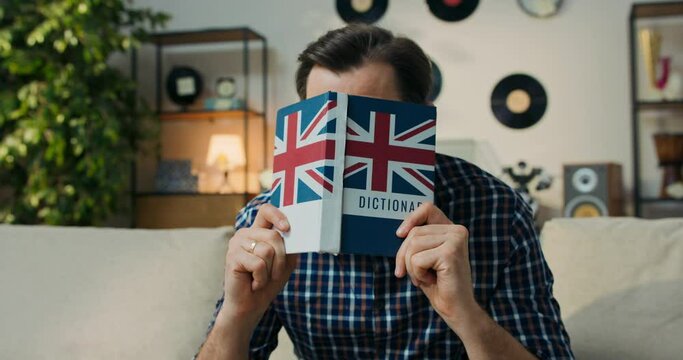 A man tries to memorize new English words. A bearded man sits on the couch and hits head with a dictionary, hoping that he will finally remember the definition.