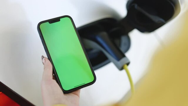 Close up green screen mock up chroma key on mobile phone. EV charging station for electric car with mobile app. Female charges batteries, uses phone with chromakey for activates start charging
