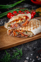 Shawarma with vegetables on a wooden board with pita sauce on a dark stone table decorated with cherry tomatoes. Fast food restaurant, delivery service.