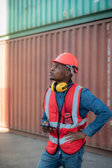 African black male worker standing, using smartphone and wearing safety work equipment clothes Work at a warehouse of Overseas shipping port, cargo, import-export industry.