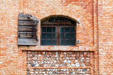 Vecchia finestra ad arco con i vetri chiusi e un vecchio  balcone di legno aperto su facciata di abitazione abbandonata in pietra e mattoni di colore rosso.