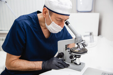 A male doctor in blue lab suit, white hat, mask and black gloves studies a stained histology slide...