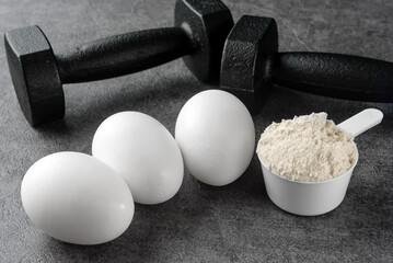The concept of sports nutrition. Dumbbells, protein scoop and chicken eggs close-up on a dark background.