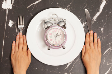Women's hands holding knife and fork, alarm clock on a plate on a marble background, top view