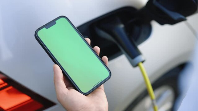 Green screen mock up chromakey display smart phone with electric car charging on street. Man charging electric vehicle with cable looking at app on mobile phone