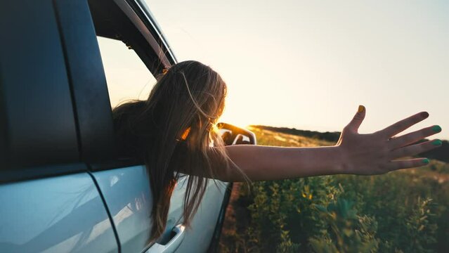 Happy family travel. Little girl leaning out of car window waving hand. Happy child girl putting her arm out of open window of a car. Summer road trip concept.