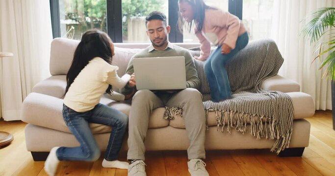 A stressed freelance father being distracted by children while typing an email. Little girls jumping on the couch and pulling man disgruntled dealing with bad behaving children in the lounge at home