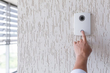 Hand of a man in a white shirt pressing the button of a door knocker on a white wall of a house, a...