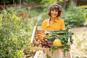 Portrait of young cheerful woman holding box full of freshly picked vegetables standing at home garden. Concept of local growing of organic food and sustainable lifestyle